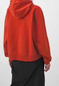 Sudadera roja con un ajuste holgado y una capucha grande; presenta puños y dobladillo acanalados; combinada con una falda negra, mostrando un look casual y en capas.