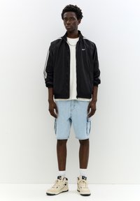 Sort zip-up jakke med hvide striber, hvid t-shirt, lys blå cargo shorts og cremefarvede sneakers med en mørk sål.