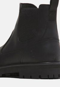 Botín Chelsea de piel negra con textura suave, paneles laterales elásticos y suela de goma con relieve que presenta un sutil detalle del logo.