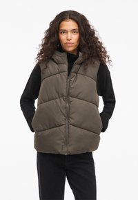 Ukošena prsluk maslinasto zelene boje, s visokim ovratnikom, prednjim patent zatvaračem i teksturom u obliku dijamantskih kvadrata. Nošen preko crne majice dugih rukava.