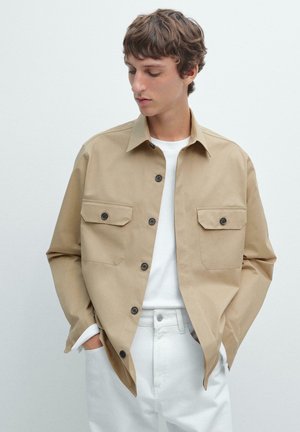Beige knapjakke med to brystlommer, lavet af et glat stof. Kombineret med en hvid t-shirt og lyse bukser.