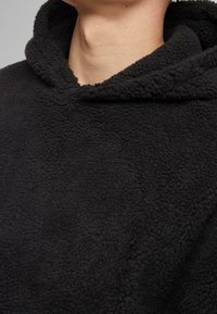 Felpa nera in pile con tessuto morbido e texturizzato, cappuccio aderente e design minimalista. Nessun logo o motivo visibile.