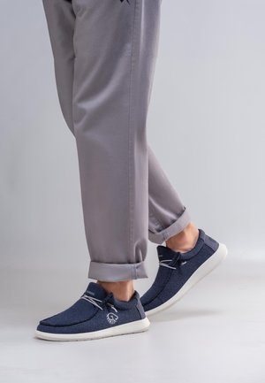 Osoba nosząca jasnoszare spodnie podwinięte na mankietach oraz granatowe, wsuwane buty sportowe z białą podeszwą i elastycznymi sznurowadłami.