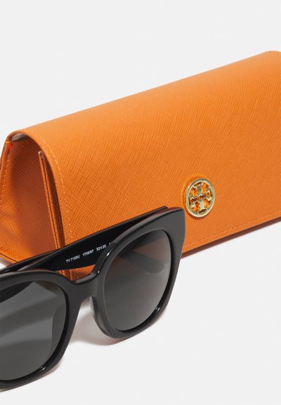 Óculos de sol pretos com lentes escuras ao lado de uma caixa laranja texturizada com um logotipo dourado na frente.