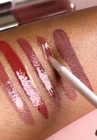 Cinque diverse tonalità di rossetti liquidi provate sulla pelle, con un pennello che applica una tonalità marrone media.