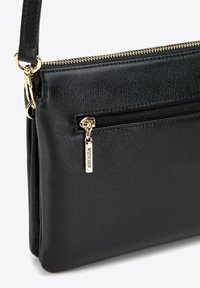 Schwarze Lederhandtasche mit glatter Textur, oben mit Reißverschluss, Seitentasche und goldfarbenen Metallakzenten. Auffälliges Markenschild angebracht.