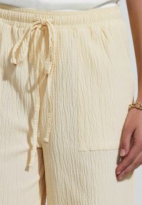 Helle gelbe Stoffhose aus kräuseligem Material mit elastischem Bund und Kordelzug, ausgestattet mit einer Seitentasche und einer strukturierten Oberfläche.