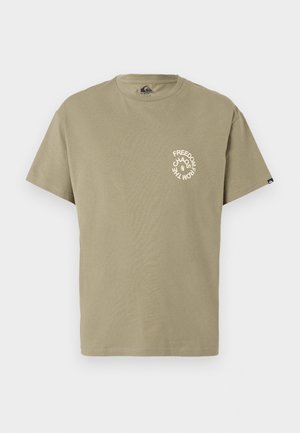 T-shirt di cotone verde oliva con scollatura tonda. Il davanti presenta un design grafico circolare bianco con il testo "LIBERTÀ DALLA CAOS." Maniche corte.