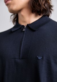 Inquadratura ravvicinata di una persona che indossa una polo blu navy con zip e un piccolo logo ricamato sul petto sinistro.