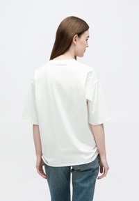 Hosszú barna hajú nő, laza fehér rövid ujjú pólót és kék farmert visel, háttal áll egy egyszínű világos háttér előtt.