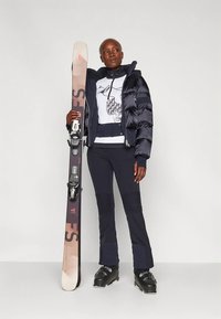 Μπουφάν πάφφλερ σε μαύρο, φορεμένο πάνω από λευκό φούτερ με γραφικό τύπωμα. Σκούρα παντελόνια με ριμπ υφή, μπότες για σκι και πέδιλα με δεσίματα.