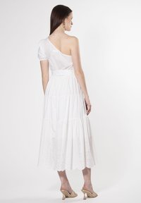 Vestido blanco de un solo hombro con manga abullonada y falda en capas. Bordado de ojales en el dobladillo. Modelo lleva tacones beige y pendientes sencillos.