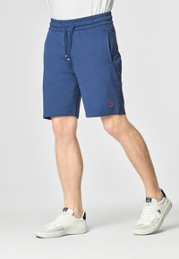 Pantaloni corti in cotone blu con vita elasticizzata e coulisse; presentano tasche laterali e un piccolo logo rosso sulla coscia sinistra. Le sneakers sono bianche.