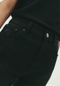 Schwarze Jeans mit hohem Bund, ausgestattet mit einem vorderen Knopfverschluss, bronzefarbenen Beschlägen und dezenten Taschenstichdetails.