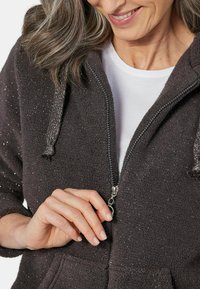 Haut à capuche en molleton gris foncé avec une fermeture éclair intégrale, des détails argentés et un cordon de serrage à la capuche. Il possède une texture douce et des poches avant.