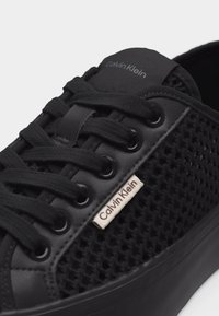 Μαύρο αθλητικό παπούτσι Calvin Klein με πλαϊνά μέρη από δίχτυ, μαύρα κορδόνια και ετικέτες με το λογότυπο στη γλώσσα και κοντά στα ρινίσματα.