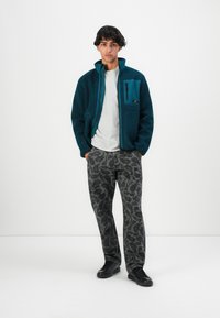 Ung mann står med hendene i lommer iført mørk teal fleecejakke, lys grå skjorte, grå bukser med pixelmønster og svarte joggesko.