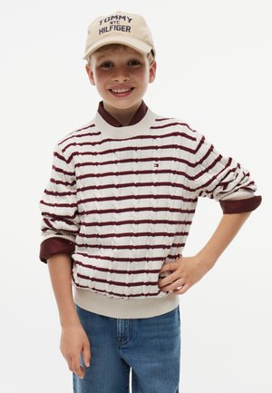 Smiling boy wearing a beige Tommy Hilfiger cap, white and maroon striped sweater over maroon shirt, and blue jeans, standing with one hand on hip.