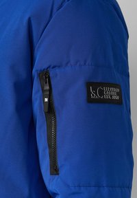 Blauer Jackenärmel mit einer schwarzen Reißverschlusstasche und einem rechteckigen Markenschild mit eingeprägtem Text. Glattes, strukturiertes Gewebe.