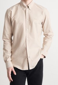 Lichtbeige overhemd met knopen, kraag, lange mouwen, witte knopen en een subtiel logo op de borst. Draag met een donkere broek.