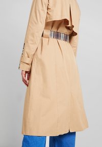 Cappotto beige con cintura a quadratini e dettagli sui polsini, indossato sopra jeans blu, visto da dietro su uno sfondo neutro.