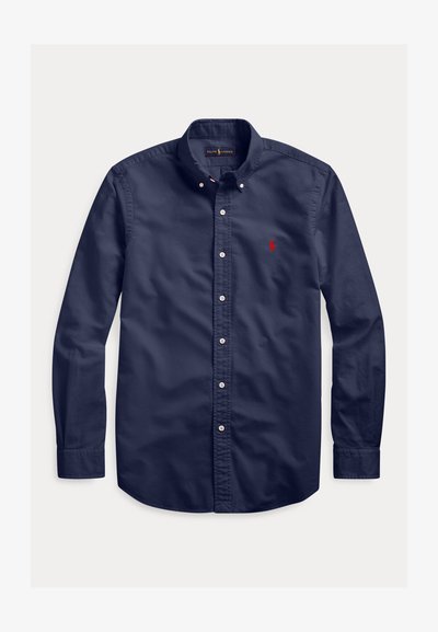 Tamsiai mėlyna ilgarankovė marškiniai su sagomis, kurių apykaklė užsegama sagomis, ir mažas raudonas siuvinėtas logotipas kairėje krūtinės pusėje.