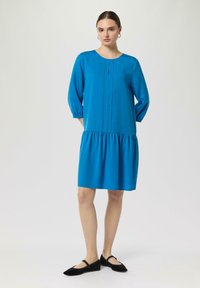 Türkises Kleid mit Dreiviertelärmeln, gefaltetem Vorderteil, rundem Ausschnitt und gerafftem Saum, kombiniert mit schwarzen Ballerinas auf einem hellen Hintergrund.