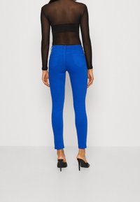 Jeans ajustados de color azul con un diseño de cintura alta, cinco bolsillos y una textura de denim suave. Se llevan con una blusa negra transparente y tacones.