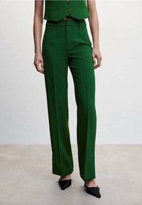Green tailored trousers with a straight leg cut, featuring front pleats and side pockets, paired with black pointed flats.