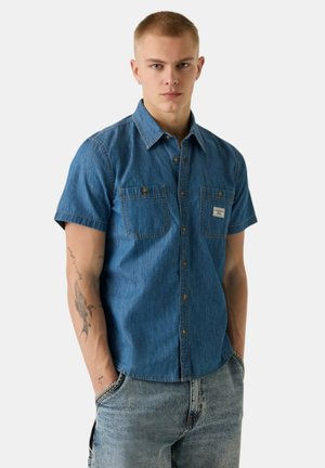 Giovane uomo con capelli corti che indossa una camicia di jeans blu a maniche corte e jeans chiari, mani in tasca, espressione neutra.