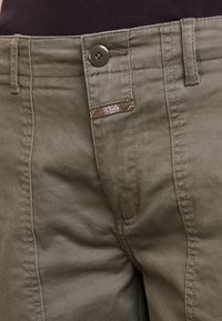 Pantalons en coton vert olive avec fermeture à bouton, couture visible et étiquette de marque près de la taille, au toucher lisse.