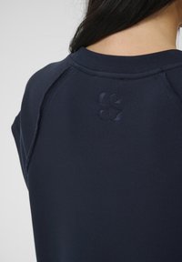 Temno modra kapuca z okroglo ovratno, kratkimi rokavi in subtilnim, vtisnjenim logotipom na hrbtu. Mehka tkanina z čistim zaključkom.