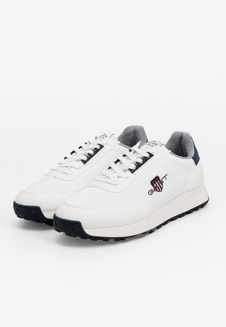 Sneaker bianche con una superficie sintetica liscia, accenti blu navy, suole testurizzate e lacci. Presentano un logo laterale e un colletto imbottito.