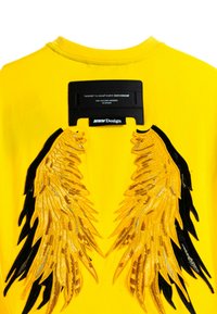 Camisa amarilla de manga corta con diseños bordados de alas negras y doradas en la espalda, con una etiqueta rectangular negra en el cuello.