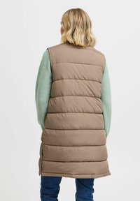 Gilet long et rembourré de couleur marron clair avec des coutures horizontales. Portez-le par-dessus un pull vert. Les fentes sur les côtés en bas facilitent le mouvement.