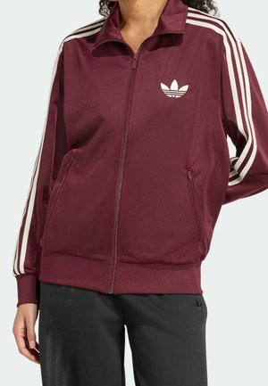 Person wearing a maroon Adidas track jacket with white stripes on sleeves and black pants with subtle stripes and logo.