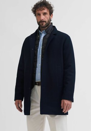 Mężczyzna z kręconymi włosami i brodą nosi warstwowy granatowy płaszcz, czarną pikowaną kamizelkę, jasnoniebieską koszulę i białe spodnie na jednolitym szarym tle.