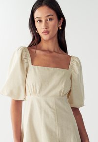 Svijetla bež haljina s puf rukavima, kvadratnim izrezom, nabranim ramenskim šavovima i oblikovanom siluetom, izrađena od teksturirane tkanine.