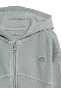 Felpa zip grigia chiara realizzata in tessuto a maglia testurizzato, con cappuccio, dettagli cuciti ai lati e un piccolo logo ricamato sul petto.