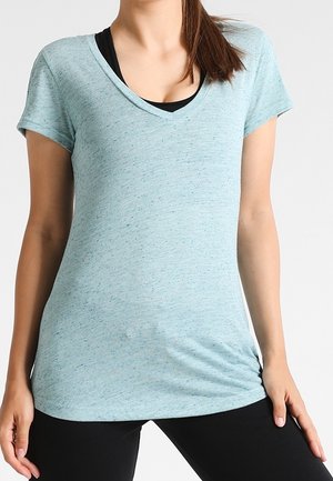 Femme portant un T-shirt bleu clair à manches courtes et col en V par-dessus un soutien-gorge de sport noir, associé à un legging noir, debout les mains le long du corps.