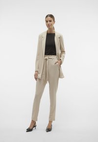 Beige op maat gemaakte blazer en bijpassende broek met een riem, gecombineerd met een zwart top. Slim fit, soepele textuur, met zwarte hakken.