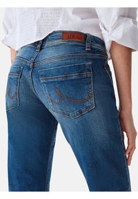 Vaqueros de mezclilla azul con un ajuste de tiro medio, que presentan una etiqueta de cuero marrón, dos bolsillos traseros con costuras curvadas y detalles de costura visibles.