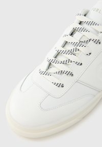 Hvit lær-sneaker med perforerte detaljer, flate hvite lisser med et svart rutemønster, og en glatt gummisåle.