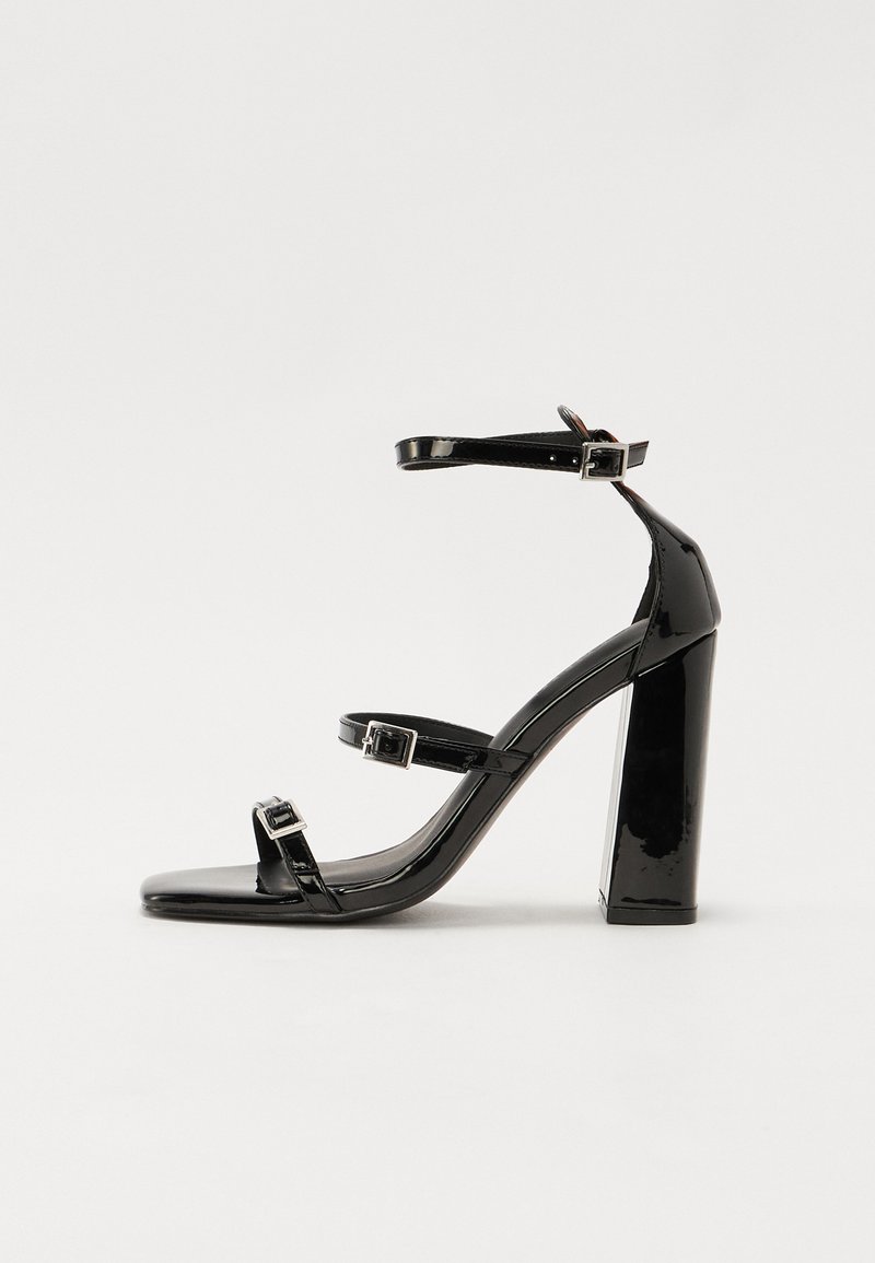 Sandales à talons vernies noires avec un bout carré, des sangles autour de la cheville et des boucles en argent. Dotées d'un talon bloc épais pour plus de stabilité.