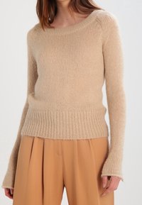 Beżowy sweter dzianinowy z dekoltem w serek, prążkowanym dołem i mankietami, charakteryzujący się miękką, puszystą teksturą oraz luźnym krojem. W połączeniu z jasnobrązowymi spodniami.