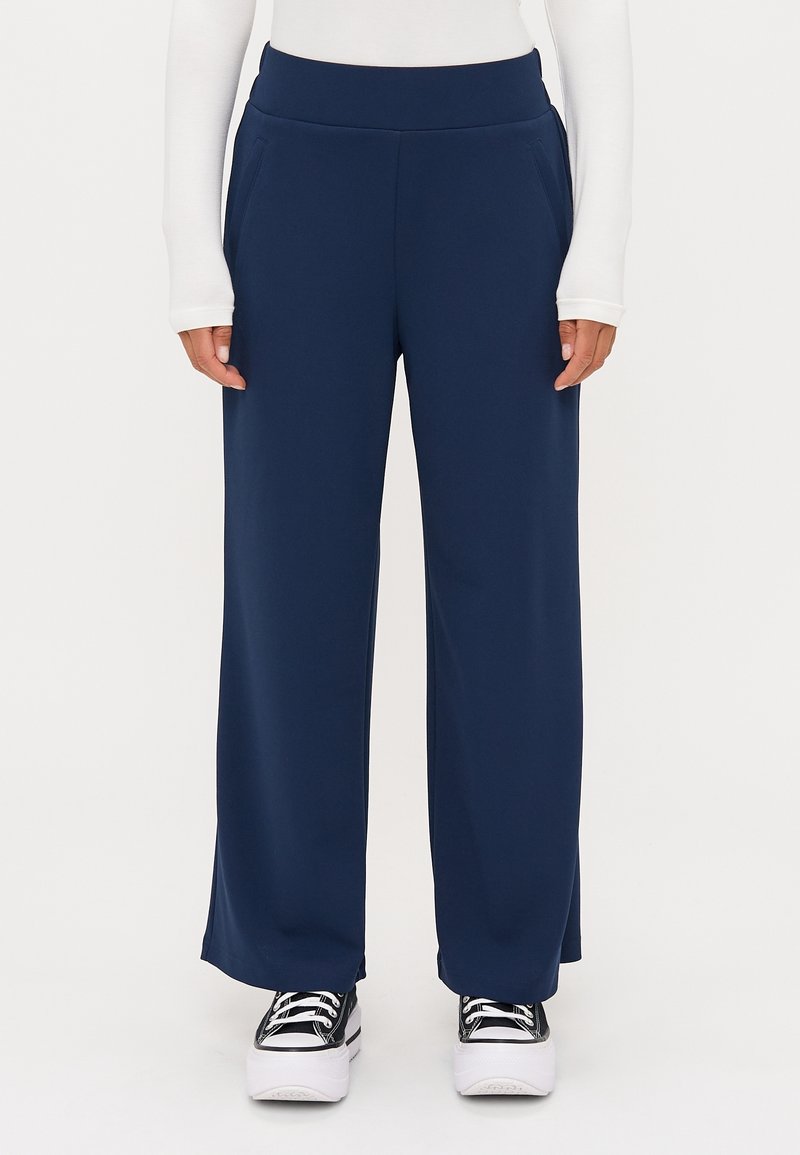 Personne portant un pantalon large bleu marine avec poches avant, un haut blanc à manches longues et des baskets à plateforme noires, debout sur un fond uni.