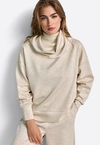 Sweatshirt bej, cu o textură moale, având un guler larg tip cowl și umeri căzuți, cu mâneci lungi și o croială lejeră, stilizat cu pantaloni asortați.