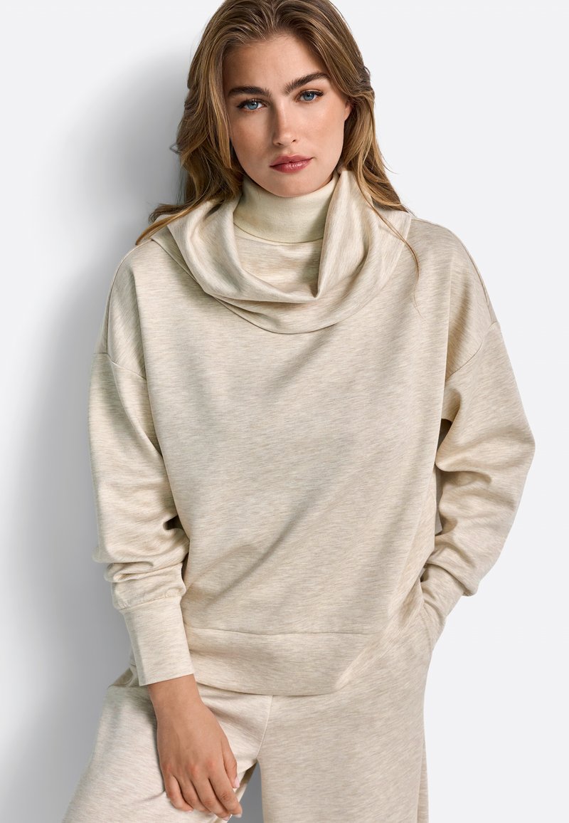 Sweatshirt bej, cu o textură moale, având un guler larg tip cowl și umeri căzuți, cu mâneci lungi și o croială lejeră, stilizat cu pantaloni asortați.