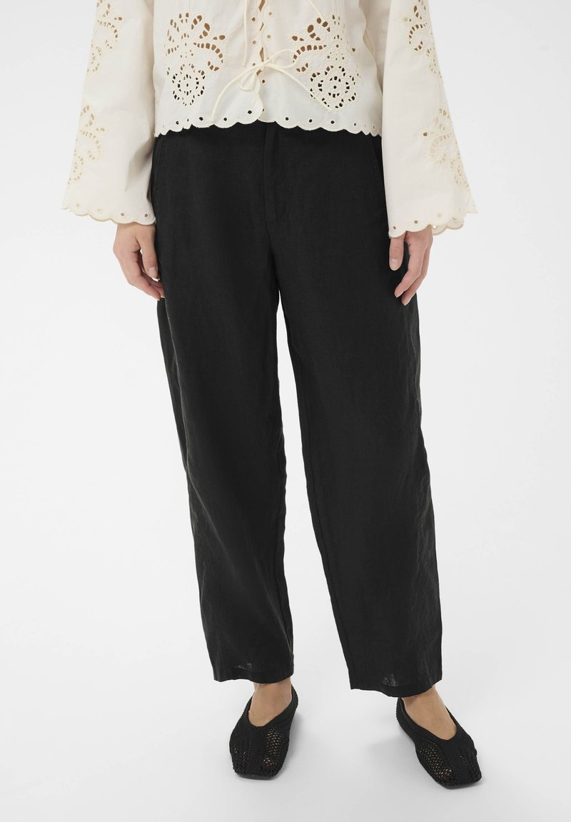 Femme portant un pantalon noir ample, un chemisier blanc à manches longues avec des motifs floraux découpés, et des chaussures noires tissées sans lacets, debout sur un sol blanc.