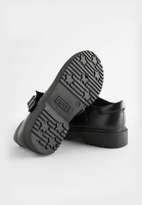 Chaussures noires avec une tige en cuir lisse, dotées d'une sangle Velcro et d'une semelle en caoutchouc texturée avec des motifs d'étoiles. Logo de la marque visible sur la semelle.
